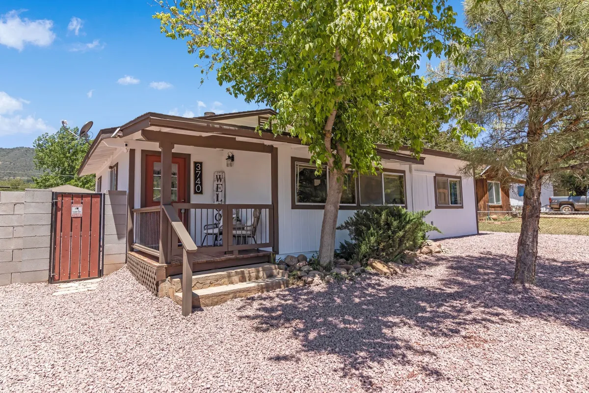 The image captures the front porch of the cabin, featuring a welcoming sign, vibrant landscaping, and a colorful door. There's outdoor seating available, inviting guests to relax and enjoy the peaceful surroundings. The porch is surrounded by greenery, connecting the home to its natural environment.