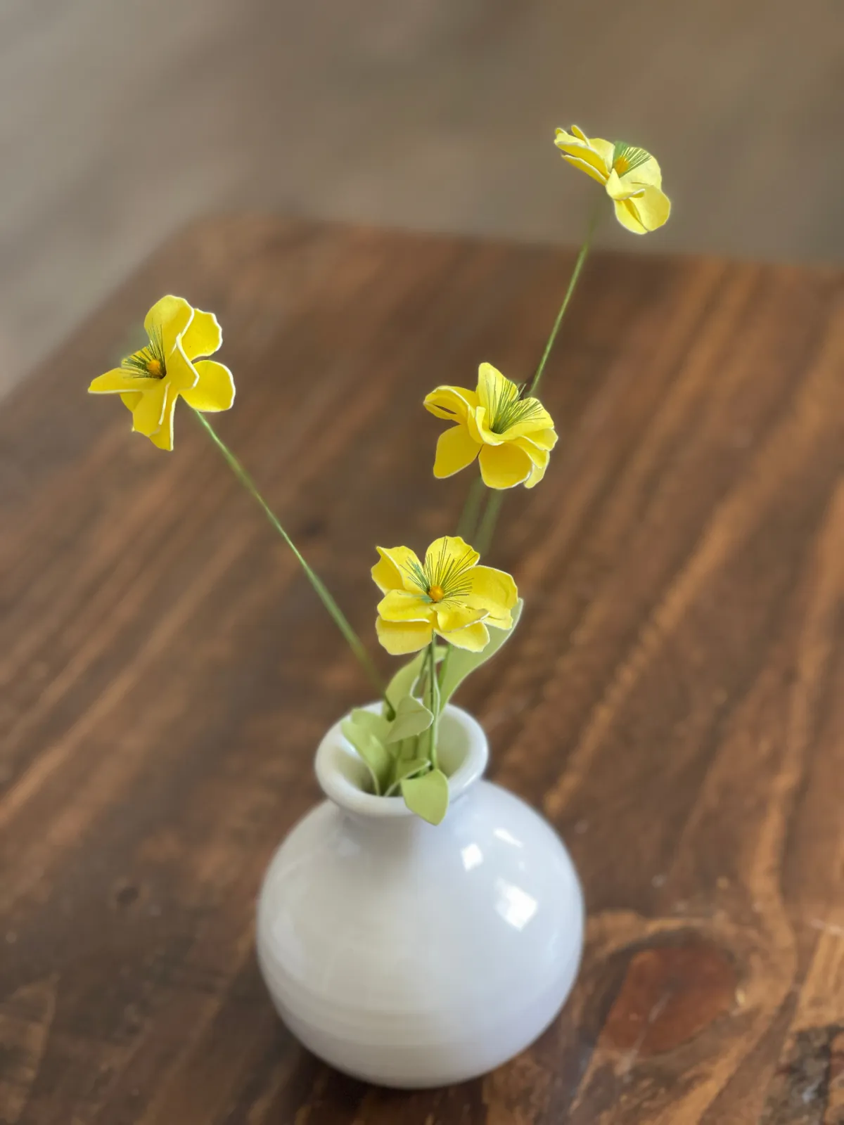 A cheerful bouquet of yellow flowers arranged in a white vase sits atop a wooden table, bathed in natural light. The fresh floral arrangement and rustic setting evoke feelings of peace and connection to nature.