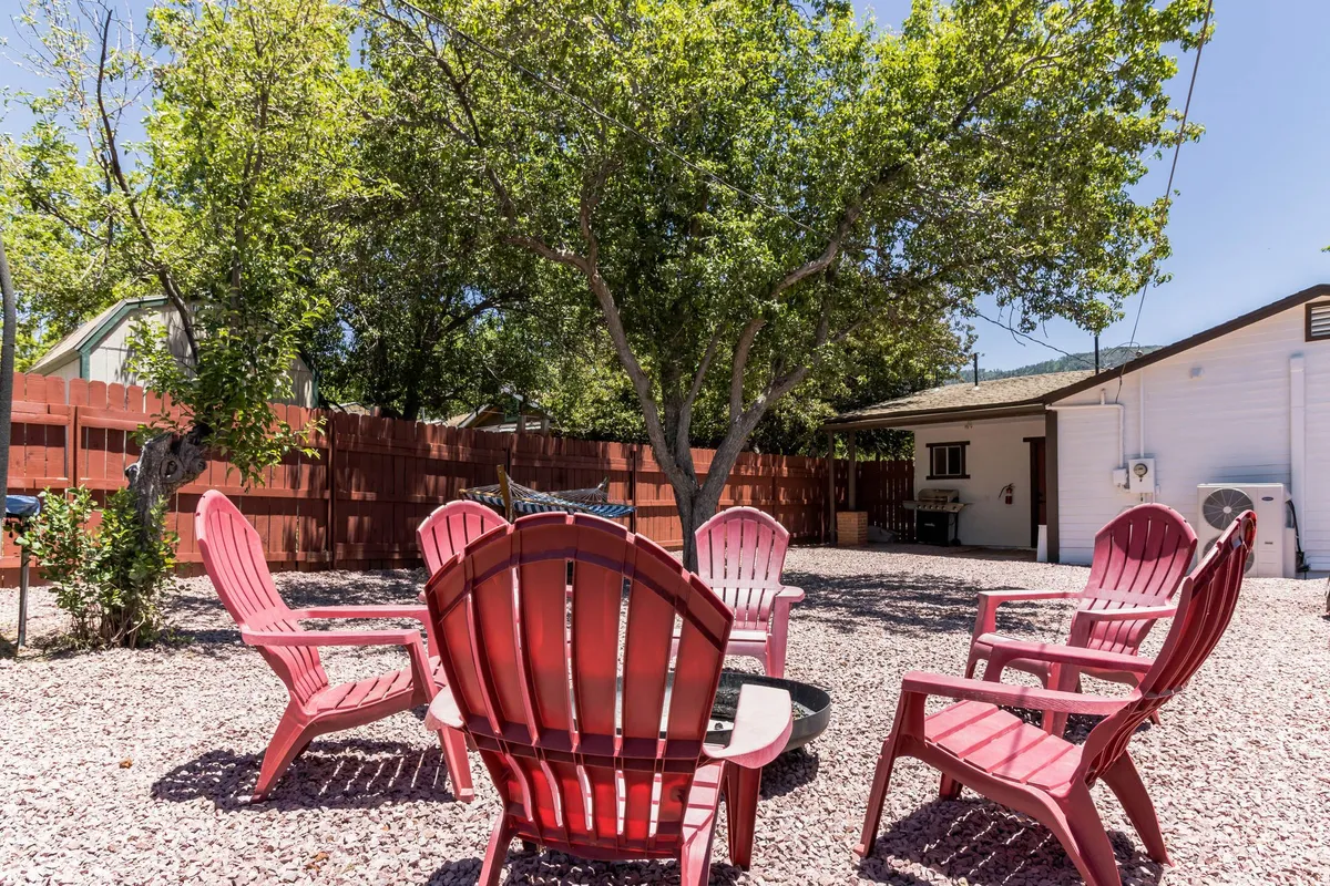 The image features red Adirondack chairs arranged beneath a large shade tree in a gravel-floored area, bordered by a wooden fence and surrounded by lush greenery. The setting is relaxed and perfect for reading or casual gatherings, emphasizing the natural and landscaped aspect of the yard.