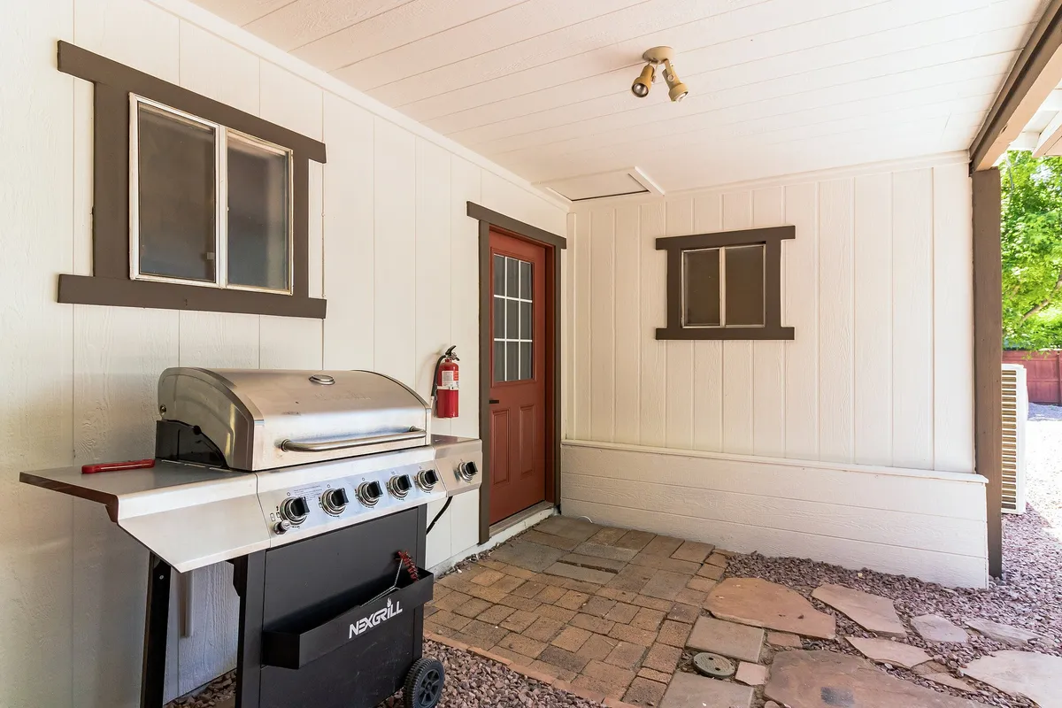 An inviting exterior scene showing the outside of the cabin, featuring a grill set up on a patio area, a striking red door, and large windows. The space is arranged for outdoor dining and barbecuing, emphasizing the potential for group gatherings after a day of adventure.