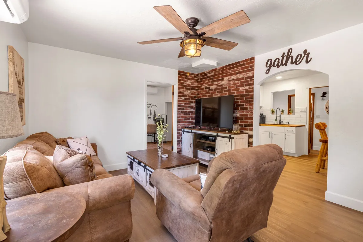A cozy living room with a comfortable sofa, sturdy coffee table, large television mounted on a brick wall, and a 'gather' sign that underscores the communal spirit of the space. The room is softly lit, inviting guests to relax, watch TV, or enjoy each other's company.
