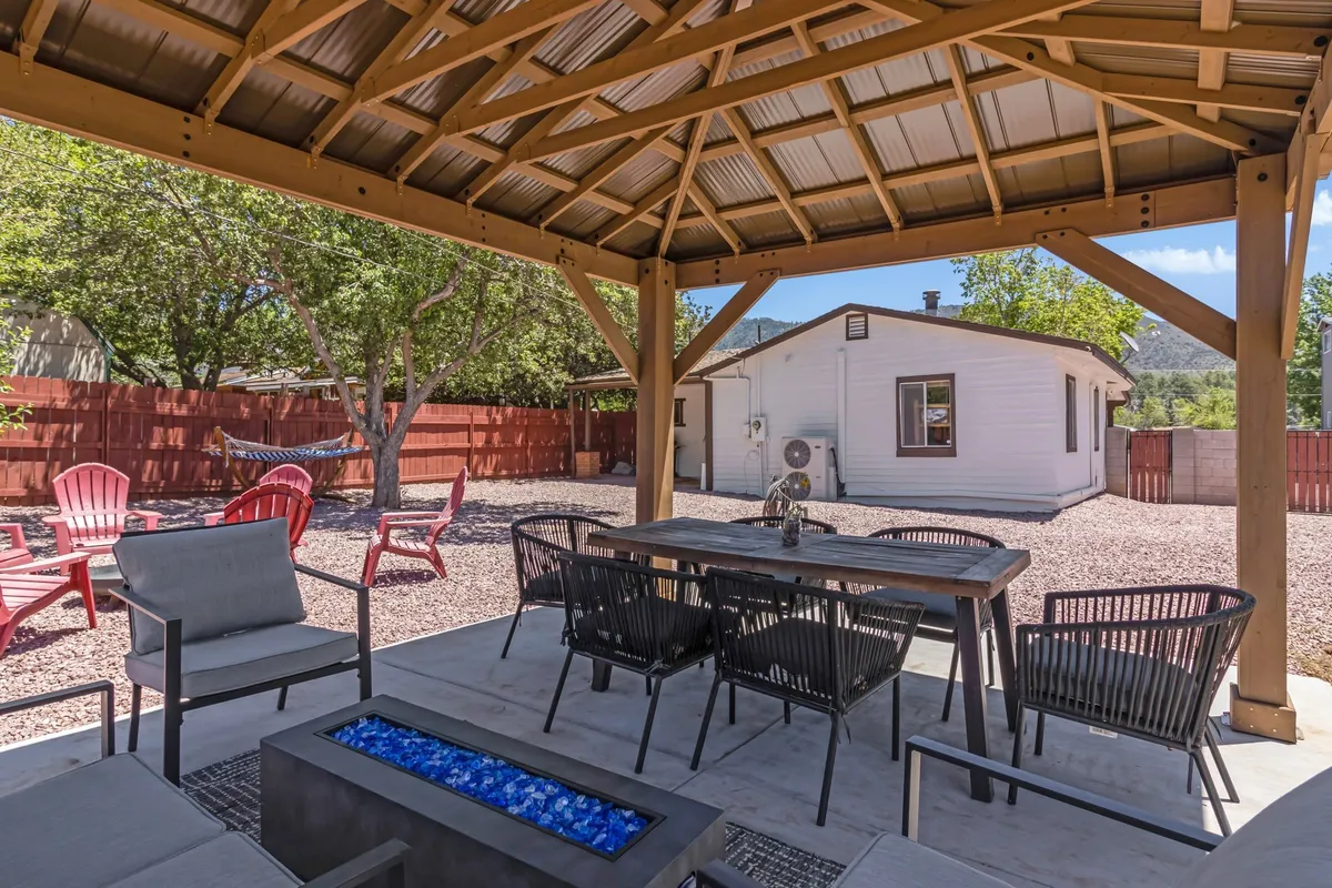 This image shows a spacious covered patio area extending into the backyard, with a dining table, a fire pit surrounded by inviting red Adirondack chairs, and mature trees providing a natural, serene backdrop. The setting is ideal for groups to dine, socialize, and relax outdoors.