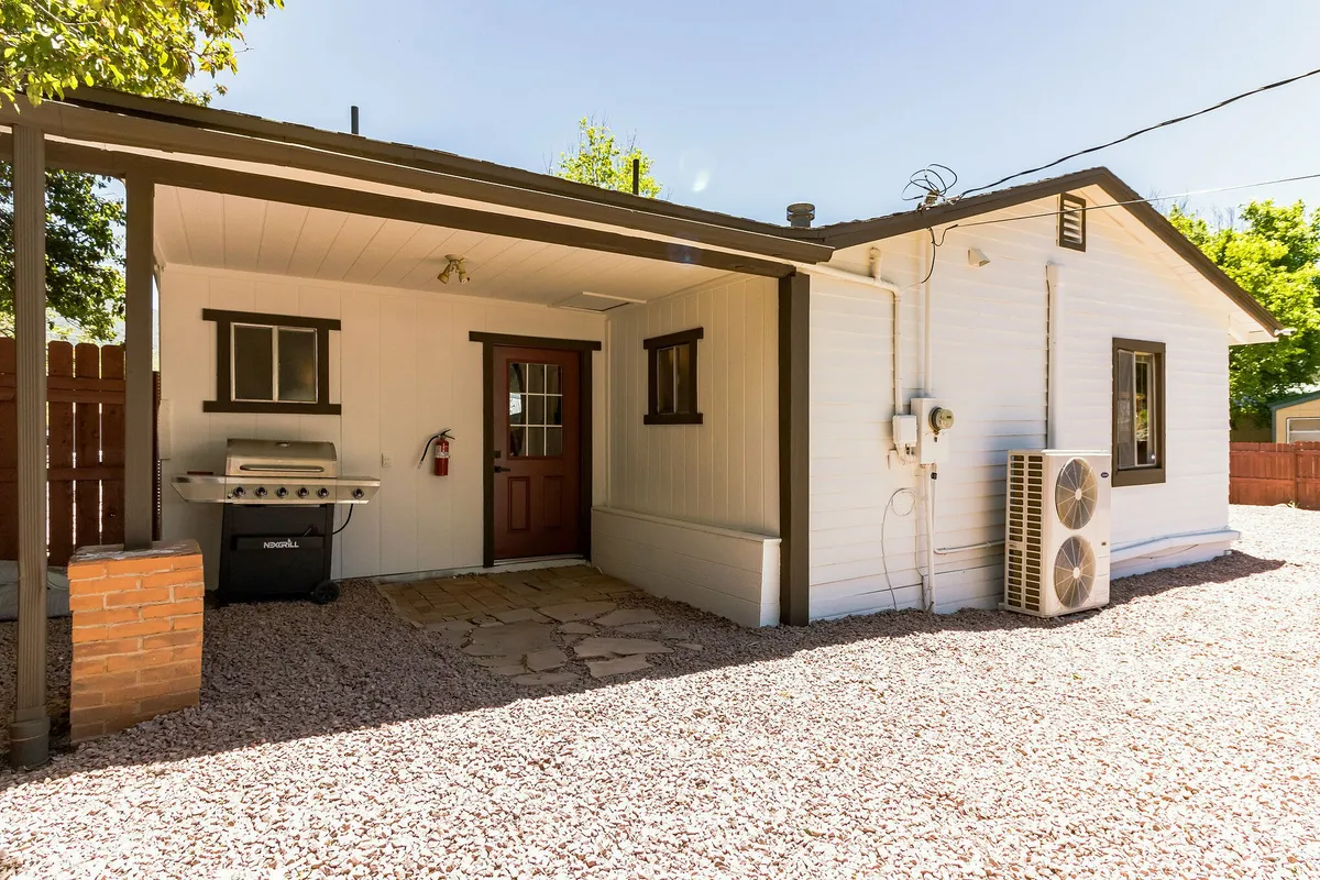 This exterior image highlights the cabin's front door, a grill, and an outdoor seating area set on a stone path. The space is bathed in sunlight, suggesting a cheerful atmosphere ideal for outdoor gatherings, barbecues, and easy access to the surrounding natural beauty.