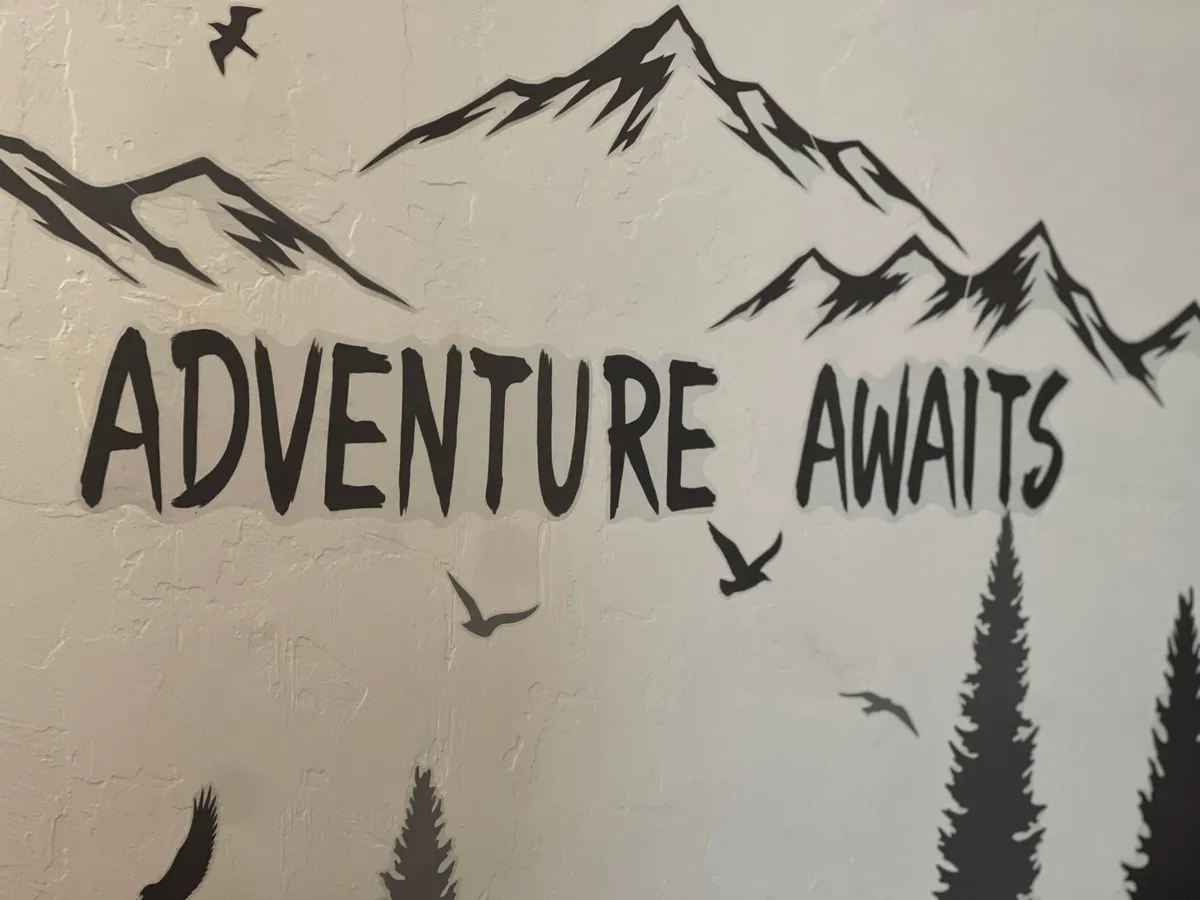 An interior scene with a bold wall art featuring the words 'ADVENTURE AWAITS,' accompanied by mountain silhouettes, birds in flight, and pine trees. The decor sets a motivational, outdoorsy mood that resonates with adventure enthusiasts.
